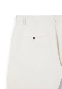 Light beige trousers with a smooth fabric texture, featuring a brown button and a back pocket with a horizontal seam detail.