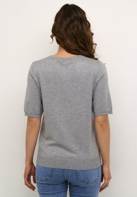 Šedý pletený pulovr s krátkým rukávem, kulatým výstřihem a žebrovaným lemem. Nesený přes modré džíny, textura vypadá hladce a lehce.