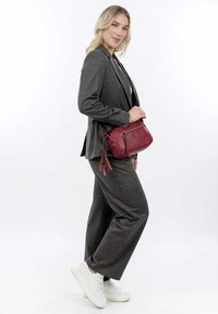 Abito grigio strutturato con blazer aderente e pantaloni a gamba larga. Il modello tiene una piccola borsa a tracolla rossa in pelle con dettagli a frange. Sneakers bianche.