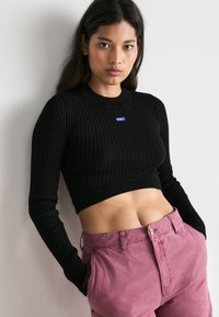 Pull noir côtelé court avec des manches longues et un col rond, arborant un petit logo bleu "HUGO". Assorti à un pantalon rose.