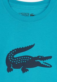 Turkusowa koszulka wykonana z bawełny z dużą czarną grafiką krokodyla z geometrycznymi akcentami. Okrągły dekolt, krótkie rękawy.