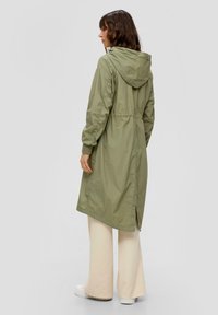 Helle olivgrüne, lange Jacke mit Kapuze, gerafftem Taillenbund, elastischen Bündchen und seitlichem Schlitz, kombiniert mit cremefarbenen, weit geschnittenen Hosen und weißen Sneakers.