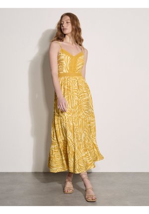 Mujer con cabello rojo que lleva un vestido largo amarillo con estampados de hojas y detalles de encaje, de pie frente a una pared lisa y clara y un suelo gris.