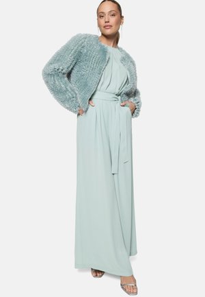 Femme portant une combinaison ceinturée bleu clair et une veste courte en fourrure texturée, debout les mains dans les poches et chaussée de sandales argentées.