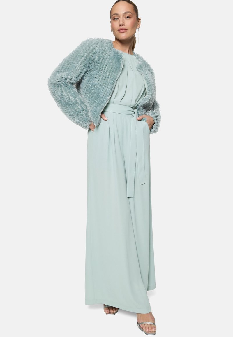 Femme portant une combinaison ceinturée bleu clair et une veste courte en fourrure texturée, debout les mains dans les poches et chaussée de sandales argentées.