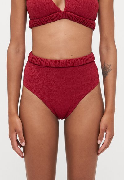 Vrouw die textuur gemaakte rode hoge taille bikini broekjes en bijpassende bikini top draagt, staand met armen relaxed langs haar zij op een effen achtergrond.