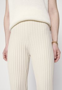 Calças de malha cor creme com riscas verticais, apresentando um cós elástico e um design ajustado. A textura parece suave e elástica.