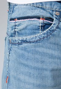 Jeans de mezclilla azul claro con una vista macro del bolsillo, con costuras contrastantes en rojo y azul, y una textura de tela natural.
