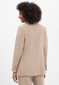 Vrouw met krullend haar, gekleed in een beige op maat gemaakt blazer en bijpassende broek, glimlachend en zijwaarts kijkend tegen een witte achtergrond.