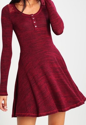 Rode jurk met lange mouwen en een uitlopende rok. Gemaakt van gestructureerde stof, met een ronde halslijn en een knoopsluiting met drie knopen. Kantaccenten op de schouders.