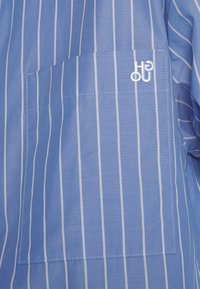 Blau-weiß gestreifter Stoff mit glatter Textur; verfügt über eine Brusttasche und ein auf der Tasche gesticktes Logo.