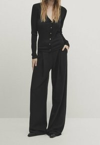 Cardigan nero a maniche lunghe con bottoni frontali abbinato a pantaloni neri a gamba larga. Entrambi i capi hanno una texture liscia e una vestibilità comoda.