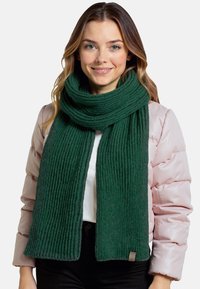 Ciemnozielony dziergany szalik o prążkowanej strukturze, noszony na jasnoróżowej kurtce puchowej. Mała metka jest widoczna na dolnej krawędzi szalika.