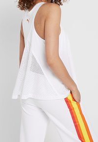 Camiseta deportiva blanca sin mangas con espalda de malla, presenta un bajo redondeado y un pequeño logo naranja. Combinada con pantalones blancos y rayas de arcoíris.