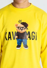 Sudadera amarilla con un oso de dibujos animados que lleva gafas, una gorra y una camisa con estampado. El texto dice "KAVAGI" debajo del diseño del oso.