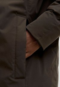 Tumši brūns žakets ar gludu, matētu tekstūru; tam ir sānu kabata un piedurkne ar aproci. Šuvju detaļa redzama šuvē.