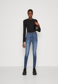 Ψηλόμεσο μπλε τζιν παντελόνι με στενή γραμμή, διαθέτει κλείσιμο με φερμουάρ και κουμπί, πέντε τσέπες και λεπτές λεπτομέρειες ραψίματος. Το μοντέλο φοράει μια μαύρη ριμπ μπλούζα.