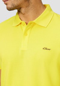 Gelbes Poloshirt aus strukturiertem Stoff mit klassischem Kragen und drei Knöpfen. Verfügt über ein kleines braunes gesticktes Logo auf der Brust.
