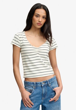 Kvinde med langt mørkt hår iført en hvid kortærmet top med sorte vandrette striber og højtaljede blå jeans med hænderne i lommerne.