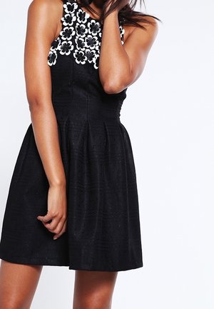 Femme portant une robe noire sans manches avec un décolleté floral blanc, debout avec une main touchant son cou et l'autre sur le côté.