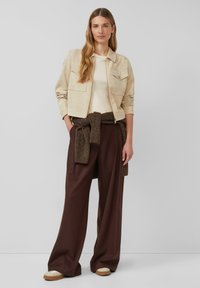 Beżowa kamizelka z zamszu z dwiema przednimi kieszeniami, kremowa bluzka, brązowe spodnie z szerokimi nogawkami z plecionym brązowym paskiem oraz beżowe sneakersy z białymi akcentami.