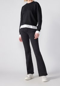 Sweat-shirt noir à coupe courte superposé sur un chemisier blanc avec des bords festonnés, associé à un jean noir évasé et des baskets blanches.