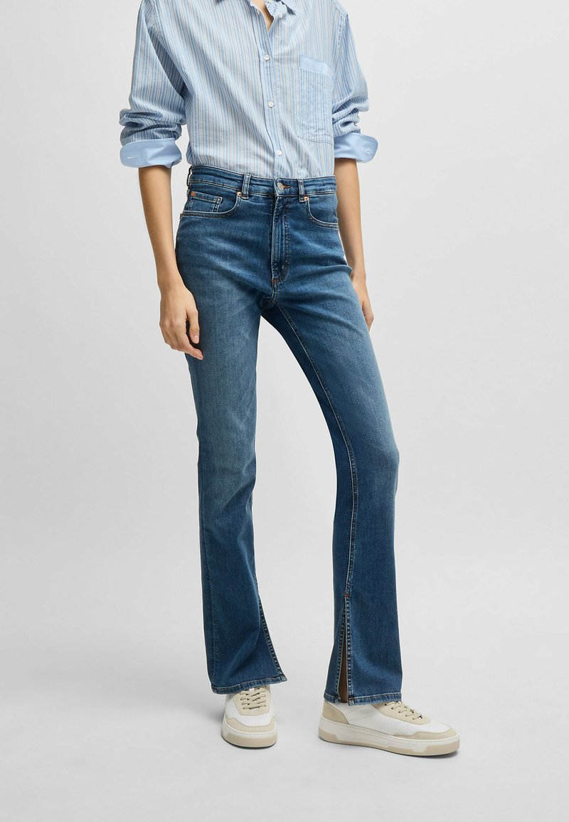 Blaue Schlagjeans mit hoher Taille, mit einem Frontreißverschluss und dezenten seitlichen Schlitzen am Saum. Getragen mit einem hellblau gestreiften Hemd und Sneakers.