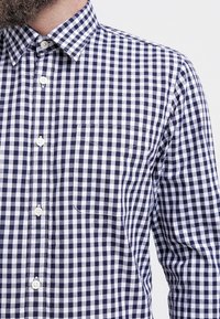 Camicia a quadretti blu navy e bianca, con colletto, maniche lunghe e una singola tasca sul petto. Il tessuto sembra di cotone e ha una consistenza texturizzata.