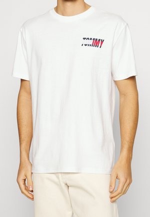 Mies pukeutuneena yksinkertaiseen valkoiseen lyhythihaiseen T-paitaan, jossa on sininen ja punainen "TOMMY"-logo vasemmalla ylärinnassa, yhdistettynä vaaleisiin beigen sävyisiin housuihin.