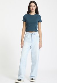 Teal crop top med korte ærmer og subtilt logo; lyseblå bredbenede jeans; sort og hvid sneakers. Glat stof, afslappet pasform, uformel stil.