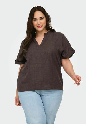 Vrouw met lang donker haar, dragend een los donkerbruin V-hals topje en lichtblauwe spijkerbroek, glimlachend met een hand licht omhoog.