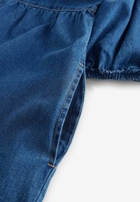 Denim tkanina v temno modri barvi, z nabranim dizajnom na dekolteju in elastičnimi manšeti. Vidno šivanje dodaja detajle.