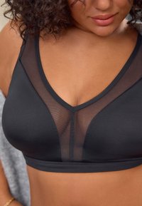 Zwarte beha met meshpanelen. V-hals, zachte stof en brede bandjes. Naaidetail in het midden voor. Gladde textuur.