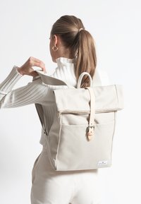 Beige kanvas ryggsäck med rulltoppdesign, läderrem och framficka. Texturerat tyg och minimaliska hårdvarudetaljer.