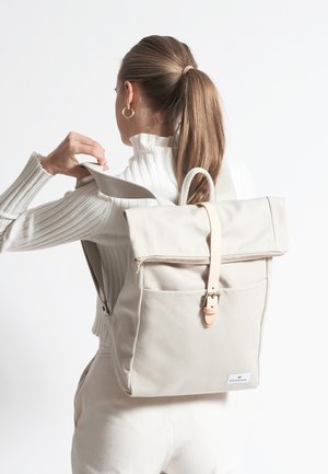 Beige Canvas-Rucksack mit Roll-Top-Design, Ledertragegriff und Fronttasche. Strukturierter Stoff und minimalistische Hardware-Details.
