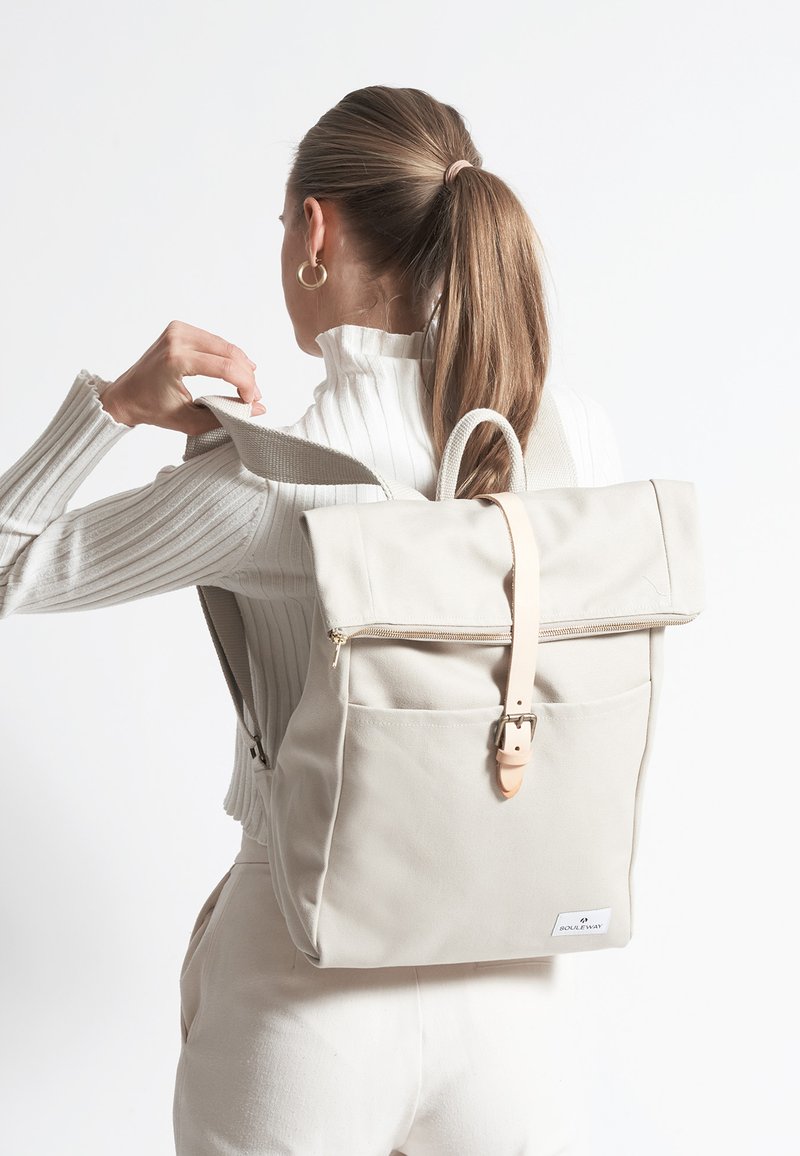 Beige kanvas ryggsäck med rulltoppdesign, läderrem och framficka. Texturerat tyg och minimaliska hårdvarudetaljer.
