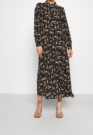 Kobieta w czarnej sukience midi z długim rękawem, z brązowo-białym liściastym wzorem, w połączeniu z brązowymi sandałami na ramiączkach i odkrytymi palcami.