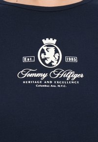 Tricou de culoare bleumarin, cu guler rotund, având un logo alb, un emblema cu leu și text: "Tommy Hilfiger Patrimoniu și Excelență" cu anul 1985.