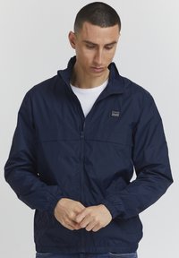 Marinblå lätt jacka i glänsande, slätt material. Har dragkedja, elastiska ärmslut och en liten logotyp på bröstet.