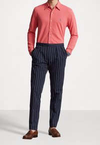 Košile s dlouhým rukávem v korálové barvě s designem na knoflíky; kalhoty s nápletem a námořnickými pruhy; hnědé kožené boty; viditelný náramkové hodinky.