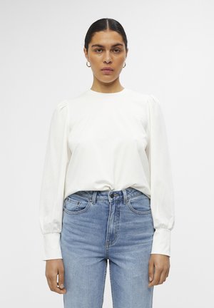 Vit långärmad topp med puffade axlar, rund halsringning och mjukt tyg, matchad med ljusblå högmidjade jeans.