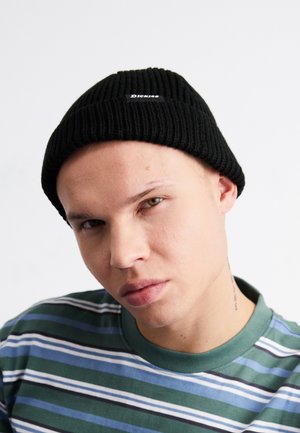 Svart ribbad mössa med en liten vit logotyp. Modellen bär en randig grön och blå T-shirt med rund halsringning.