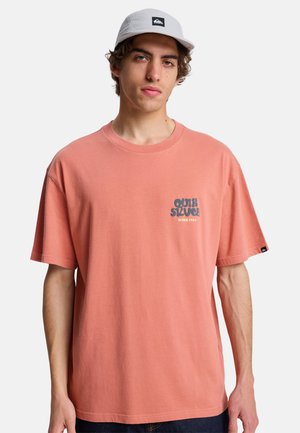 Junger Mann trägt ein korallenrotes Quiksilver-T-Shirt und eine weiße Kappe, steht vor einem schlichten hellgrauen Hintergrund.