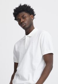 Vit polo-shirt i bomull med klassisk krage, korta ärmar och en diskret logotypdetalj. Tyget har en mjuk, texturerad yta.