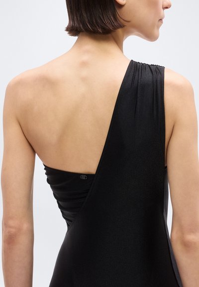 Γυναίκα με κοντά μαλλιά που φοράει ένα μαύρο φόρεμα με έναν ώμο, δείχνοντας την πλάτη και το δεξί της χέρι μπροστά σε απλό ανοιχτό φόντο.