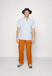 Jaunas vyras dėvi juodą plačios skrybėlės kepurę, šviesiai mėlyną marškinėlį, oranžines kelnes ir pilkus sportinius batelius, stovėdamas priešais paprastą baltą foną.