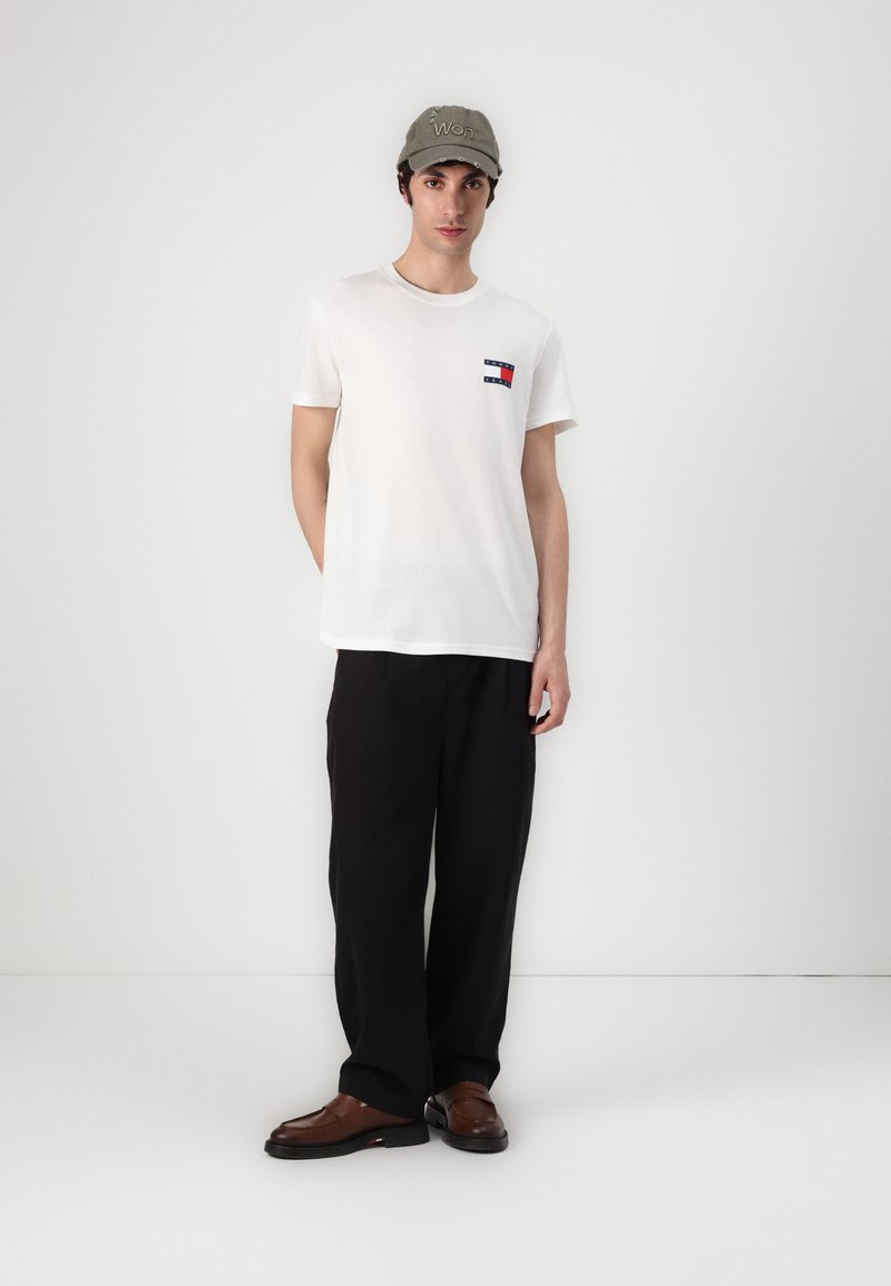 Witte t-shirt met een klein rood en blauw logo-patch, gecombineerd met zwarte wijde broek en bruin loafers. Model draagt ook een groene pet.