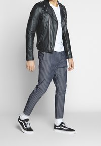 Svart läderbikerjacka, vit t-shirt, grå randiga byxor och svarta sneakers med vita sulor, stående mot en neutral bakgrund.