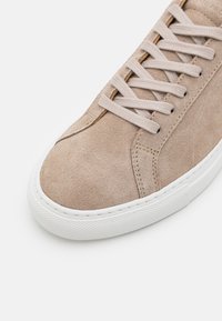 Baskets en daim beige avec lacets beiges et semelle en caoutchouc blanche, gros plan montrant la texture et les détails des coutures sur le bout et le côté.