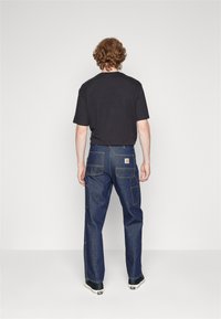 Dark t-shirt paired with blue denim pants featuring multiple pockets, visible stitching, and a branded label on the back. Black shoes.